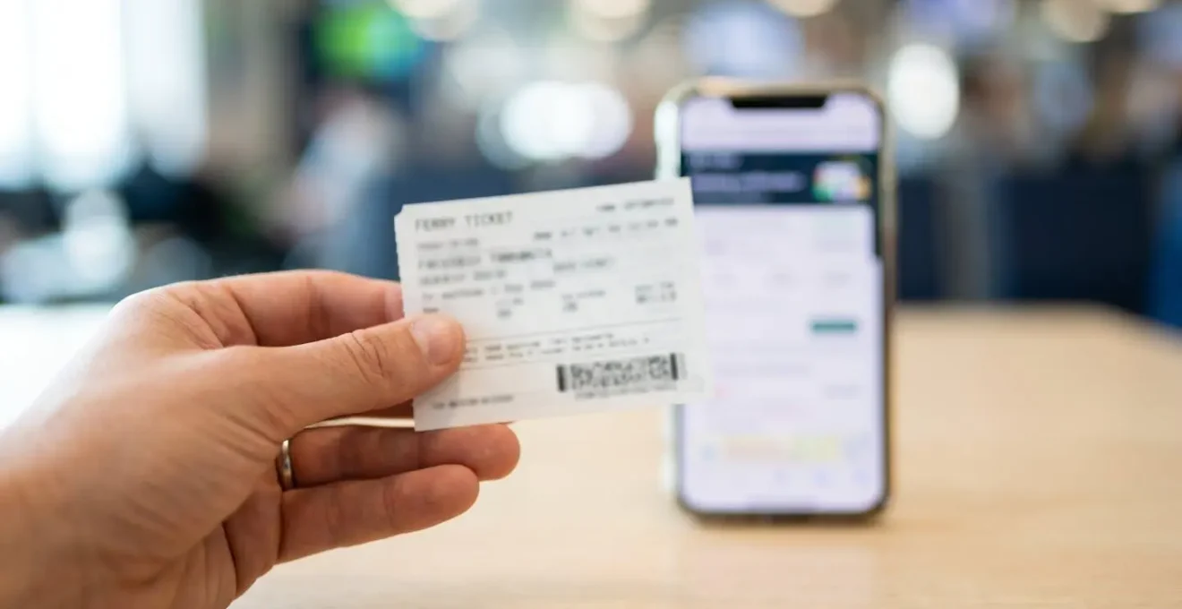 Gros plan sur une main tenant un billet de ferry avec un smartphone affichant une page de réservation en arrière-plan, texte flouté